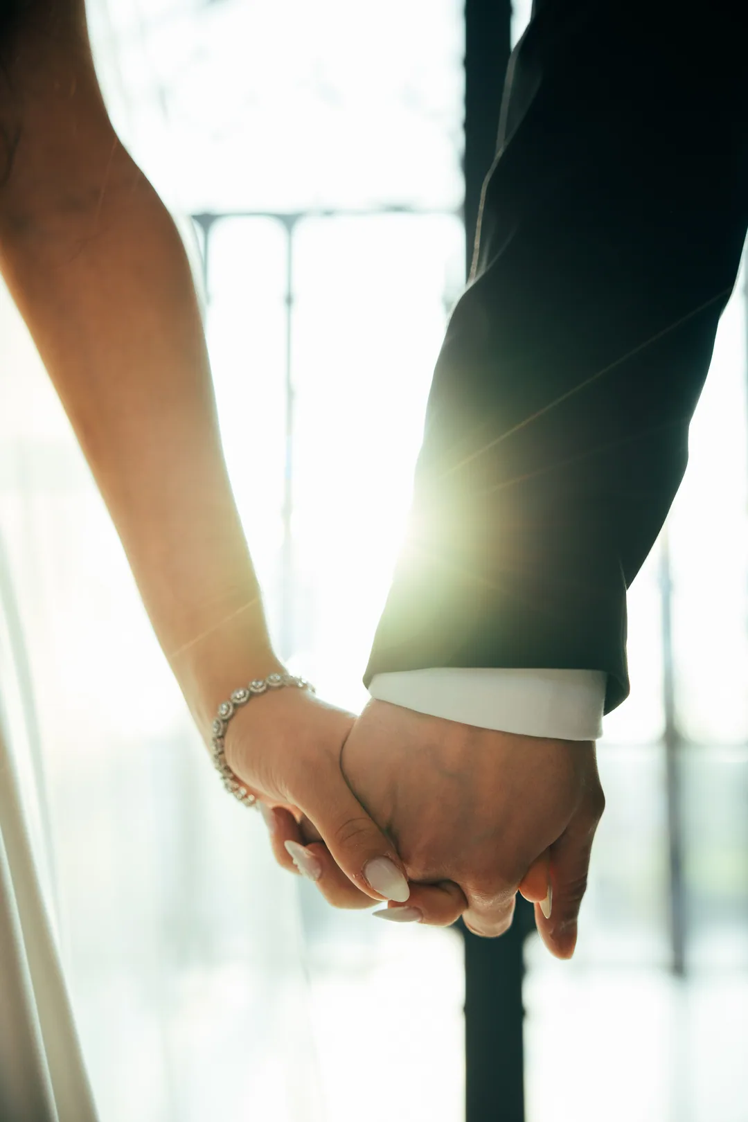 The couple's clasped hands against the window light, her bracelet catching the sun
