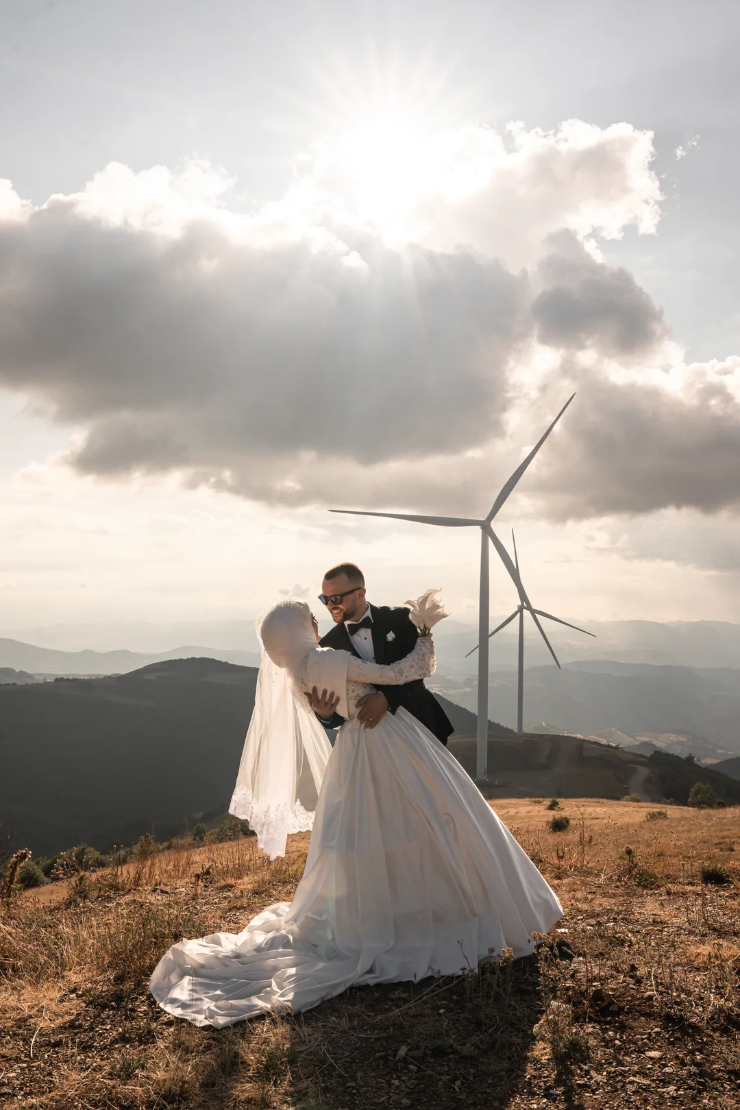 Groom kissing the bride on a mountain ridge, wind turbines behind and the sun breaking through the clouds