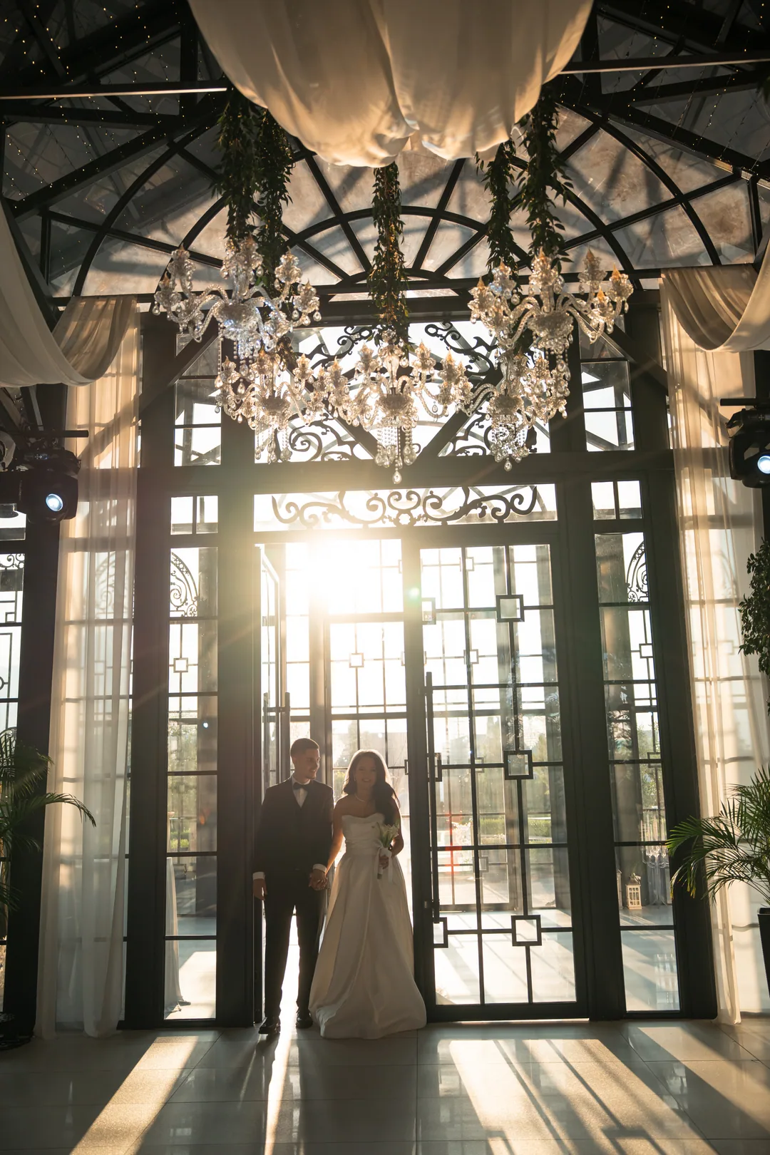 Couple silhouetted hand in hand against sunlit glass doors under a crystal chandelier