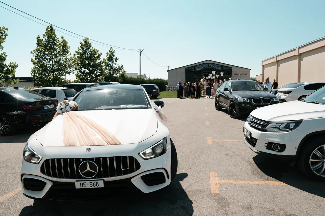 White wedding car with a pink ribbon across the bonnet, guests gathered by the venue